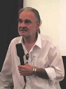 An unshaven middle-aged white man with grey hair is shown from the waist up, looking to his right. He is wearing a plain white shirt with darkened glasses hanging from the neck, and in his right hand, flanked by a silver bracelet, he holds a large black microphone.