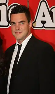 A dark brunette man in a black suit smiles at the camera