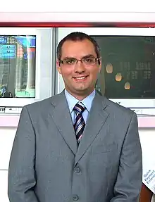 A photograph of a man with dark hair and a cleft chin wearing glasses, a grey suit, and a colourful tie while looking at the viewer and smiling