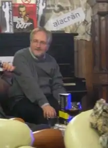 a bearded man with glasses, wearing a grey jumper, sitting on a sofa