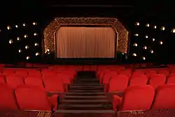 interior shot of red velvet seats facing a gilded decorated proscenium arch flanked by decorative wall lights