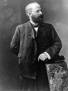 Eduard Bernstein standing next to a chair and looking rightwards. He is resting his hand on the chair. (from Socialism)