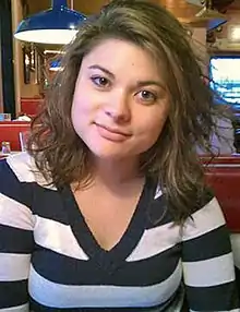 A Caucasian woman with shoulder length dark hair and brown eyes, lit from just above the frame, wearing a black and white striped top with a V-neck cut, looks at the camera with her head tilted slightly. Behind her is the interior of a restaurant.