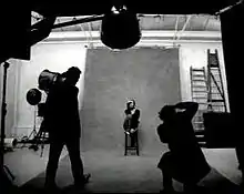 The black-and-white portrait of a brunnete woman, who is sitting in a chair. She is wearing a black sweater and jeans. In front of her, two men, one standing up, and the second taking photos are observable.