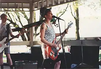 A rock band performing live. To the left, a woman plays a white guitar and in the centre, a woman plays a red bass.