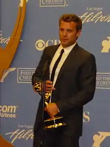 A man with dark hair, wearing a  black suit, including a black tie and white T-shirt also holding a gold statuette