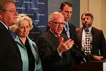 Barr gestures his right arm as his left hand rests on a podium, with two people on each side