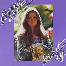 The album cover for Give It Up. It features a photo of Bonnie Raitt with her arms crossed. The photo is atop a purple background. The words "BONNIE RAITT" and "GIVE IT UP" are written in cursive, and are positioned at a slanted angle in the top-left and bottom-right corners of the cover respectively.