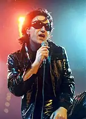 A man with black hair, black sunglasses, and a black leather attire speaks into a microphone.