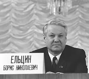 A man sitting at a desk with a tag with Russian text on it.