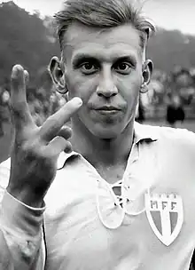 A blonde man raises his right hand to the camera with three fingers up; he is dressed in a light-coloured football kit.