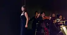 Soprano Megan Maria Hart is standing in front of the Bourbon Baroque period instrument orchestra. Violinist Nicolas Fortin has raised his bow, harpsichordist John Austin Clark's hands are hovering above the keys.