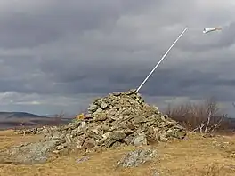 A pile of stones in a grassy clearing in late autumn. A pole with a small windsock protrudes from its right