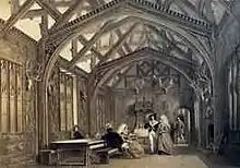 A large room with an arched roof, with the timbers visible. Some people are sitting at a table, and others are standing.