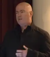 A close-up image of bald man, who is looking to the left of the camera while speaking at a conference.