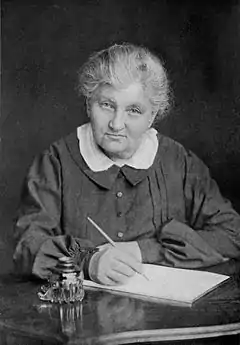 A grandmotherly woman of about 70 is seated, pen in hand, at a desk with a writing pad.