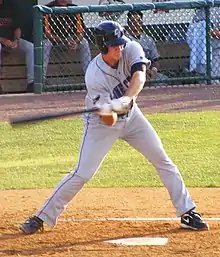 A man in a gray baseball uniform emblazoned with blue letters reading "CONNECTICUT" swings a baseball bat right handed, checking his swing.