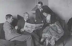 Black-and-white photograph of three men in suits and one woman seated in a room and looking at an open newspaper