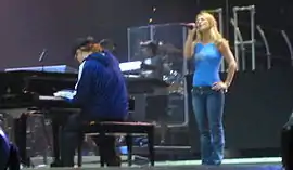 Image of a blond female performer. She is wearing blue jeans and a sleeveless light blue top. She is standing next to a piano player, who is playing a big black piano. Her left hand is in her hip while her right hand holds her mic while she is singing. Two members of the crew stand a few meters behind them.