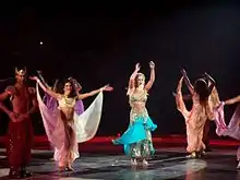 A blond female performer. She is wearing a Bollywood-inspired outfit, while surrounded by a group of dancers.