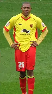 A young man wearing a yellow shirt, red shorts and red socks, standing on a grass field