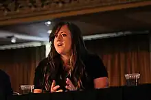 White woman with long brown hair in a black short sleeved shirt talking and gesturing with both hands in a large room