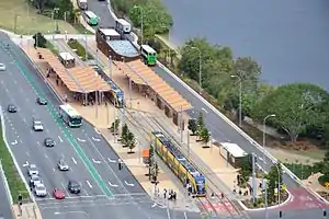 An aerial view of the station and bus interchange. The station is a single track station with platforms on either side. There are street-level open-air structures featuring passenger canopies on both platforms. Bus stops and roads are visible either side of the station.