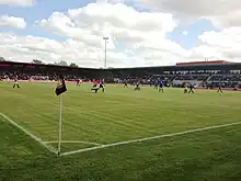 A football match is ongoing between two teams. A corner flag is in the foreground, facing two football stands full of people. The sky is lightly cloudy and an unlit floodlight is visible in the background.