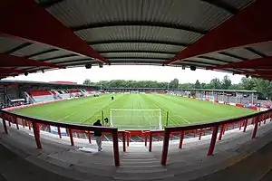 A view of a football pitch from behind the goals. There is a stand with red, white and black seating to the left. There are covered stands without terracing on the right and beyond the pitch. In the forefront is terracing with red barriers and a roof above. Green trees can be seen beyond the ground.