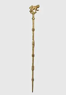 A gold staff topped with a sculpture of a cat and a mouse attempting to crawl inside the cat's bag.