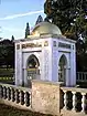 Roughly cubic white stone structure, topped by a gilded dome. Each of the four faces of the structure features a large pointed archway, and the two arches facing the camera also feature ornate Arabic calligraphy.