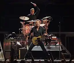 A man in dark clothing standing on a stage while holding a guitar in front of a crowd. Behind him is a man with his arm raised, holding a drum stick and sitting behind a drum set.