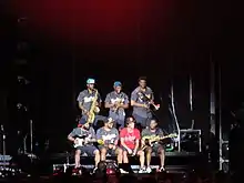 A group of seven men sit and stand on a stage against a dark backdrop.