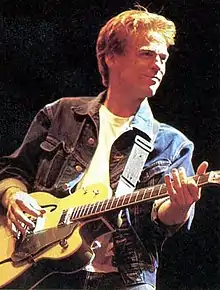 Man in a denim jacket and white shirt playing a guitar