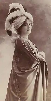 A white woman, standing, wearing a costume that includes a turban with multiple feather plumes.