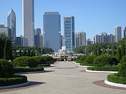 Path with hedges on either side, with a fountain and skyscrapers in the distance.