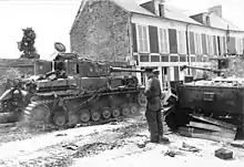 Two tanks are positioned on either side of a street with a soldier standing in the middle of the road; the road is damaged and scattered with debris.