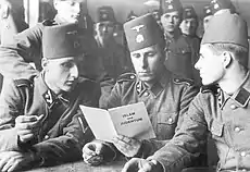 three soldiers in SS uniform and wearing fez headgear reading a pamphlet