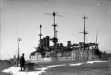 A large ship, badly rusting, lies tied up alongside a pier. Long trestles to carry small boats have been installed along the length of the deck on both sides of the ship