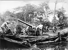 An impressive artillery piece is pulled across a small stream. Men in African colonial-style dress stand around.