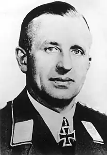 The head of a man, shown in semi-profile. He wears a military uniform with a military decoration in shape of an iron cross displayed at the front of his shirt collar. His hair is dark and combed to back.