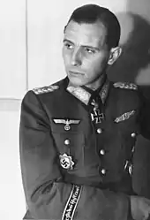 Black-and-white portrait of a man in semi profile wearing a military uniform with various military decorations, his dark hair is parted and combed back.