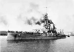 A large gray battleship with a tall tripod mast sits in harbor