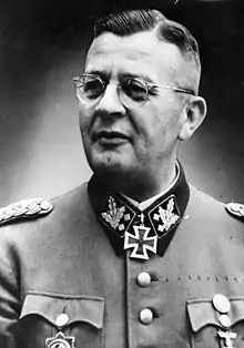 A man wearing a military uniform, glasses and neck order, in the shape of a cross. He has short hair that is combed back and a determined facial expression.