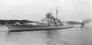 A large light gray warship bristling with guns sits in harbor