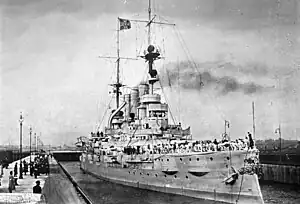 A large gray battleship sits in dock, crew members in white uniforms crowd the ship's deck