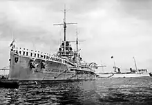 A large battleship, with sailors lining the side rails, sits in harbor. A large white ship passes behind.