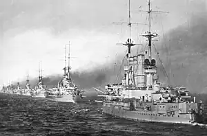 Several gray warships steam in line, thick smoke pours from each ship, darkening the sky