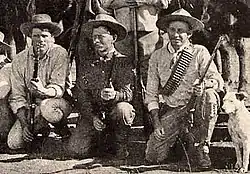 Photo taken in 1893 of three Bulawayo field scouts kneeling in front of their horses. Bob Bain on the left, Burnham in the middle, Maurice Gifford and his dog on the right.  Burnham is dressed in his Arizona clothes and is holding his Winchester model 1873 .44WCF rifle