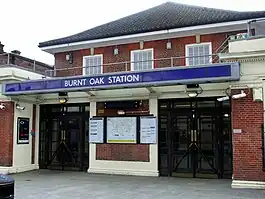 A red-bricked building with a blue sign reading "BURNT OAK STATION" in white letters and four surveillance cameras in front of the doors